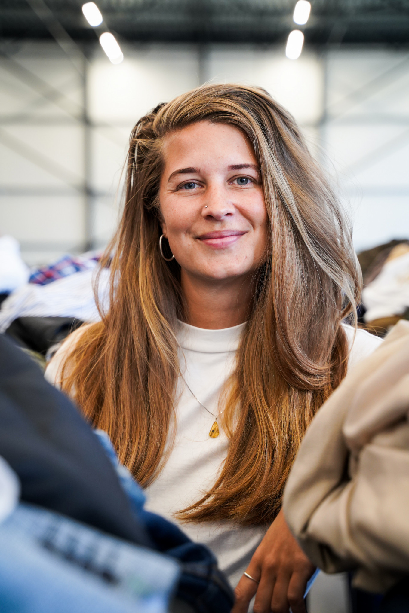 Een witte vrouw van rond de 30 jaar met lang donkerblond haar en een wit t-shirt lacht in de camera. Ze staat tussen bakken met kleding in het sorteercentrum van het Leger des Heils. 