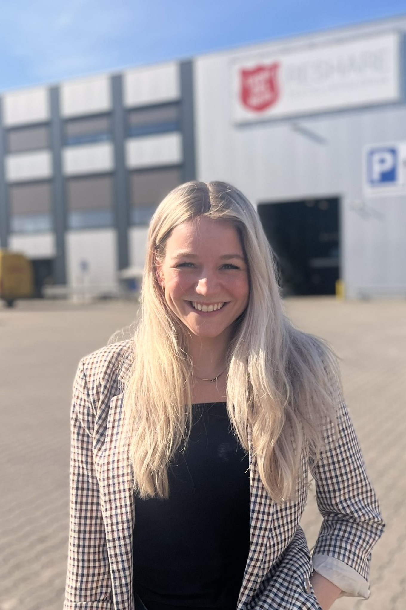  Een witte, jonge vrouw met blond haar lacht breed in de camera. Ze draagt een witte blouse en een zandkleurige colbert. Ze wordt omgeven door blauwe containers met kleding en textiel er in.