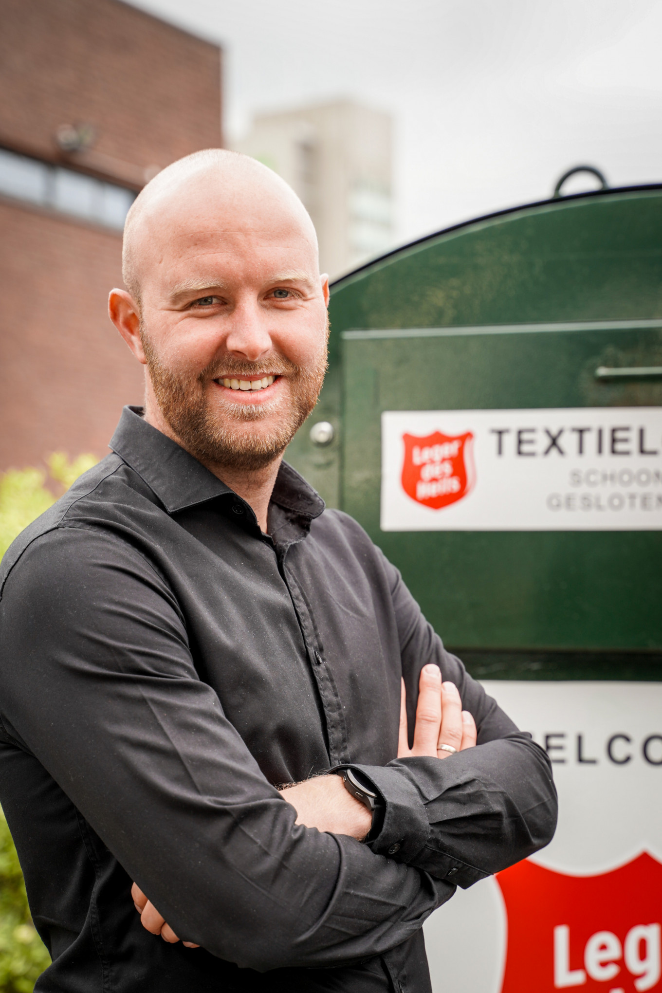 Een witte man van rond de 35 jaar met een kleine baard en een kaal hoofd en een zwart overhemd aan, staat met zijn armen over elkaar voor een textielcontainer van het Leger des Heils. Hij lacht vriendelijk.