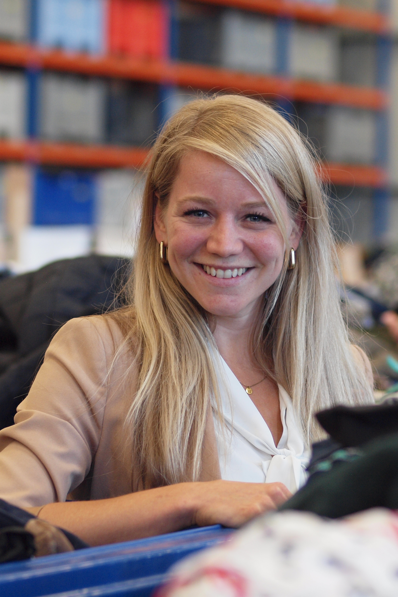  Een witte, jonge vrouw met blond haar lacht breed in de camera. Ze draagt een witte blouse en een zandkleurige colbert. Ze wordt omgeven door blauwe containers met kleding en textiel er in.