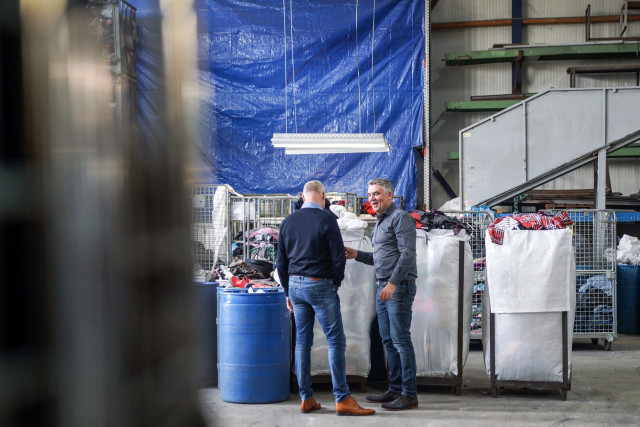 Twee mannen staan in een loods, naast een paar bakken kleding, met elkaar te praten.